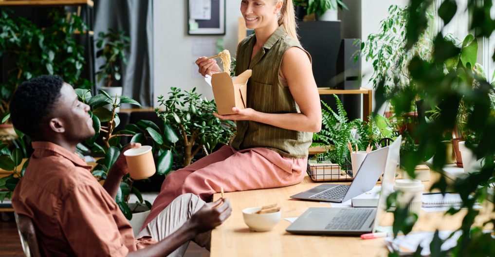Young smiling female office manager eating wok and talking to colleague Young smiling female office manager eating wok and talking to African American male colleague sitting by workplace among green plants