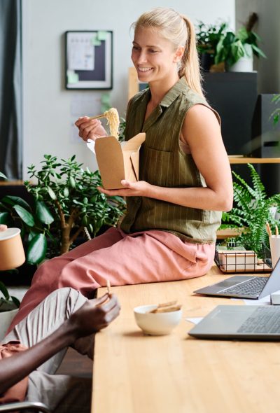 Young smiling female office manager eating wok and talking to colleague Young smiling female office manager eating wok and talking to African American male colleague sitting by workplace among green plants