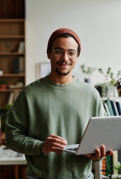 Young confident IT engineer in casualwear using laptop Young confident IT engineer in casualwear looking at camera in office while using laptop and working with online or coded data