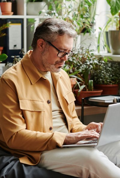 Serious mature businessman in casualwear and eyeglasses networking in office Serious mature businessman in casualwear and eyeglasses with laptop on knees networking in office while sitting on soft bag chair